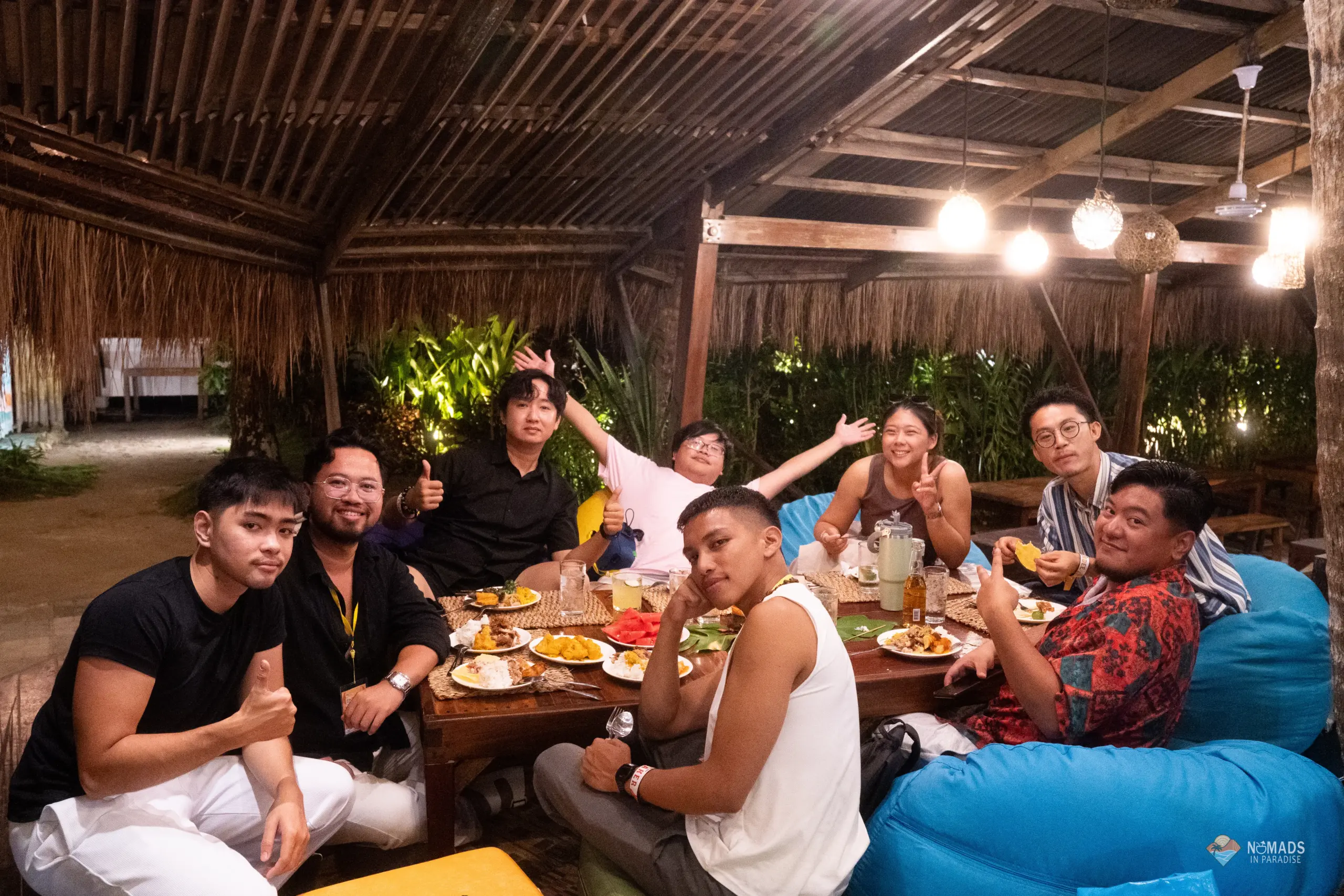 Eight cheerful Nomads in Paradise attendees dining on beanbags under thatched roof