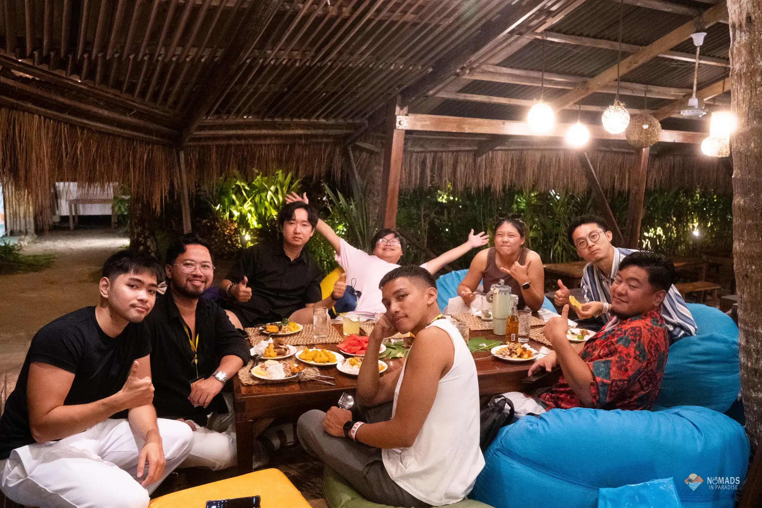 Communal outdoor dinner group photo — diverse attendees sharing meal under tropical pavilion