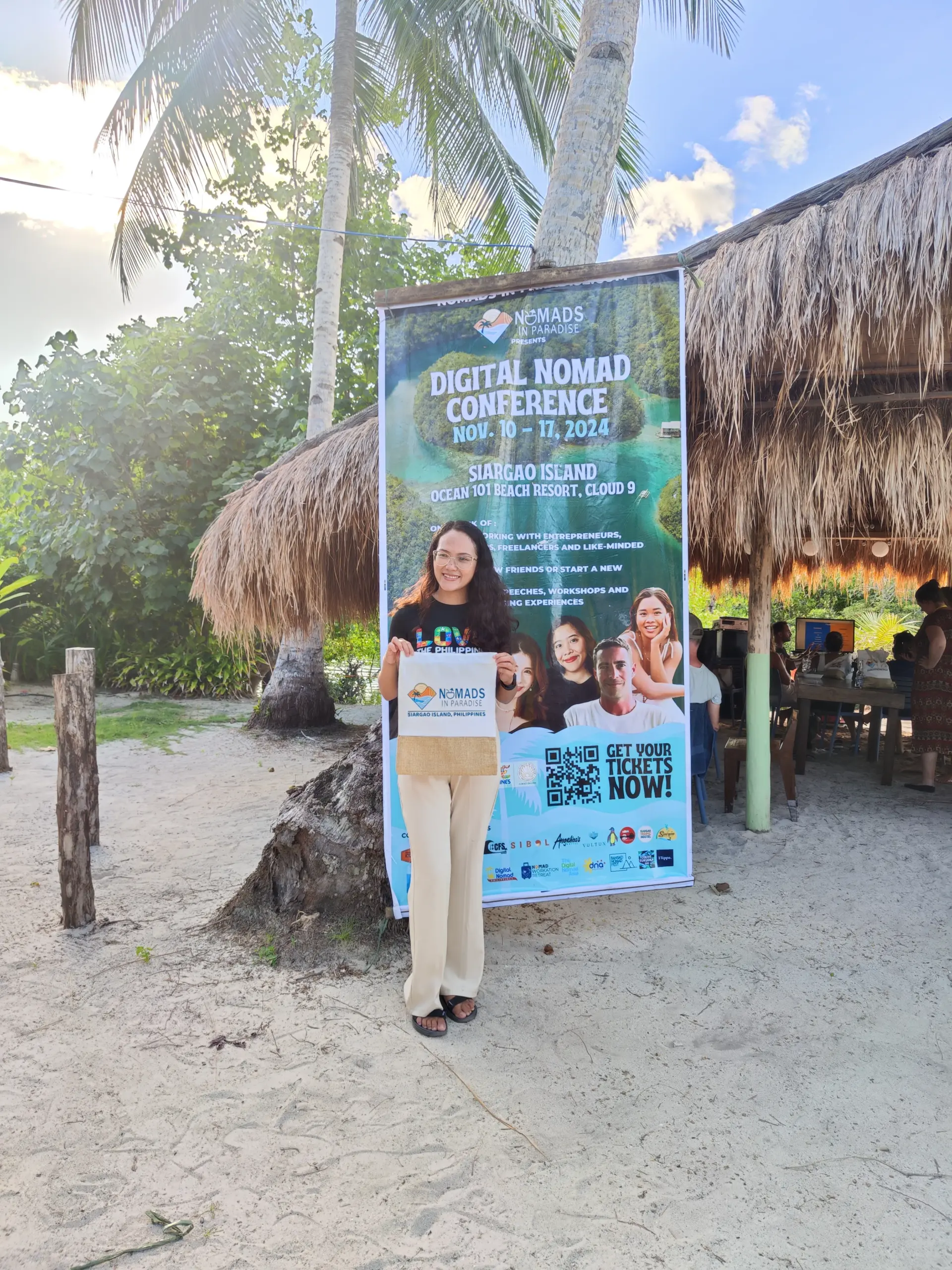Woman holding Nomads in Paradise tote bag in front of Digital Nomad Conference ticket banner