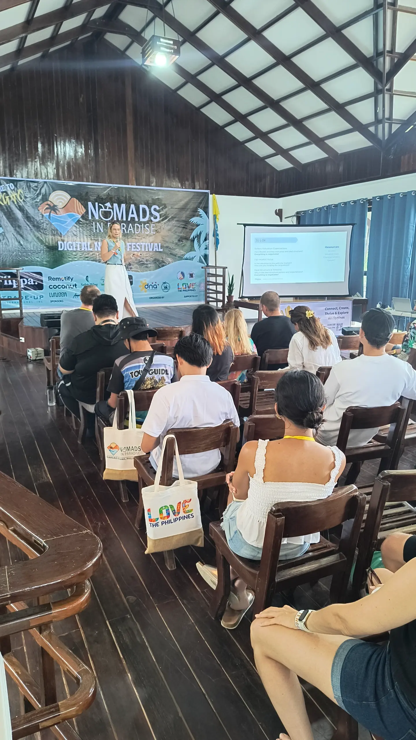 Female speaker presenting to seated attendees in wooden conference hall at Nomads in Paradise