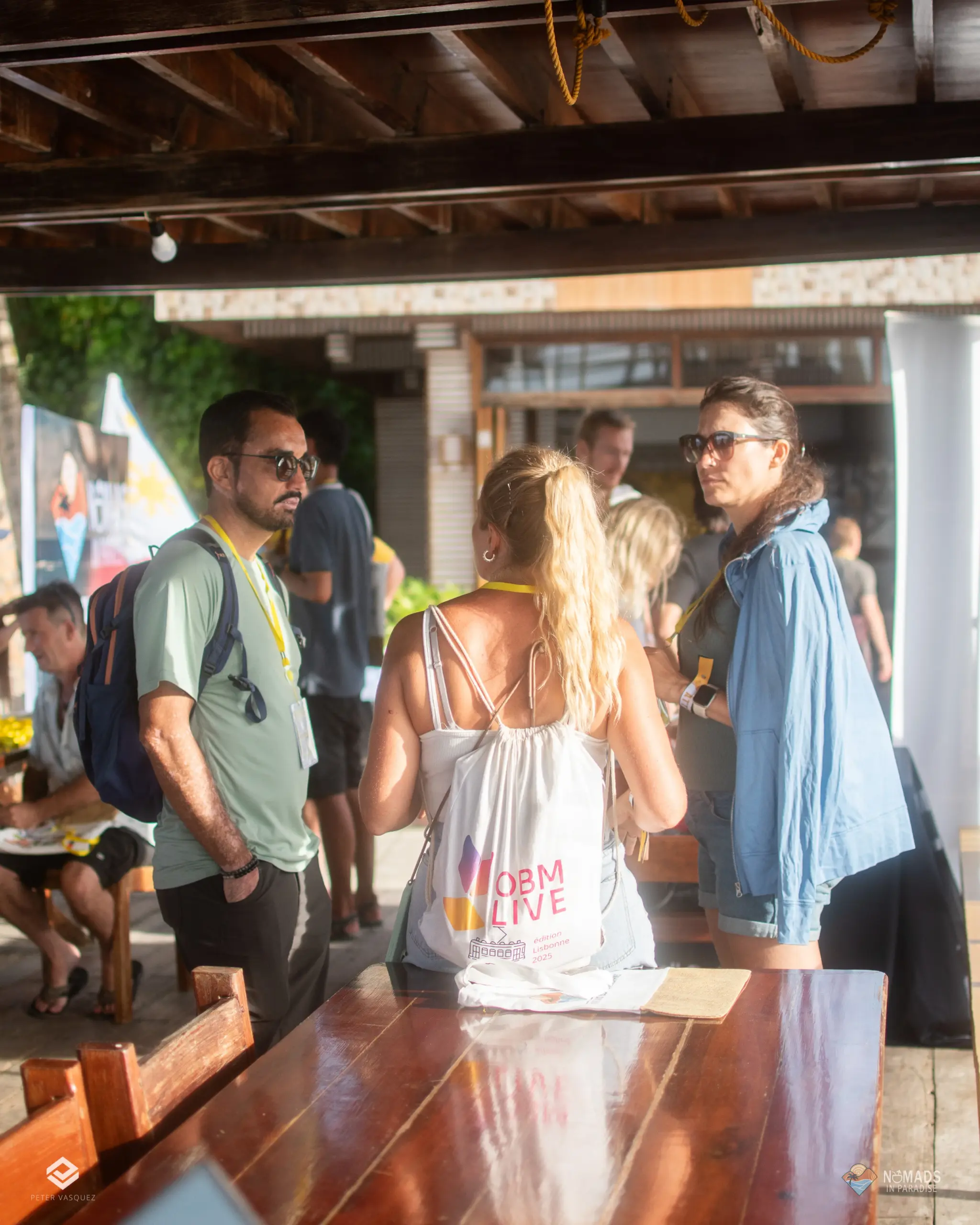 Three attendees with yellow lanyards chatting under wooden pavilion — casual networking day one