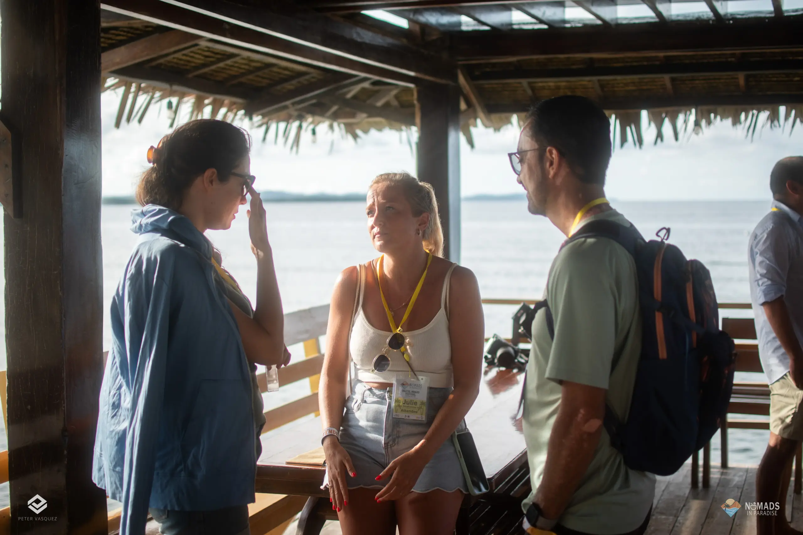 Three attendees chatting under thatched deck overlooking calm ocean at Nomads in Paradise