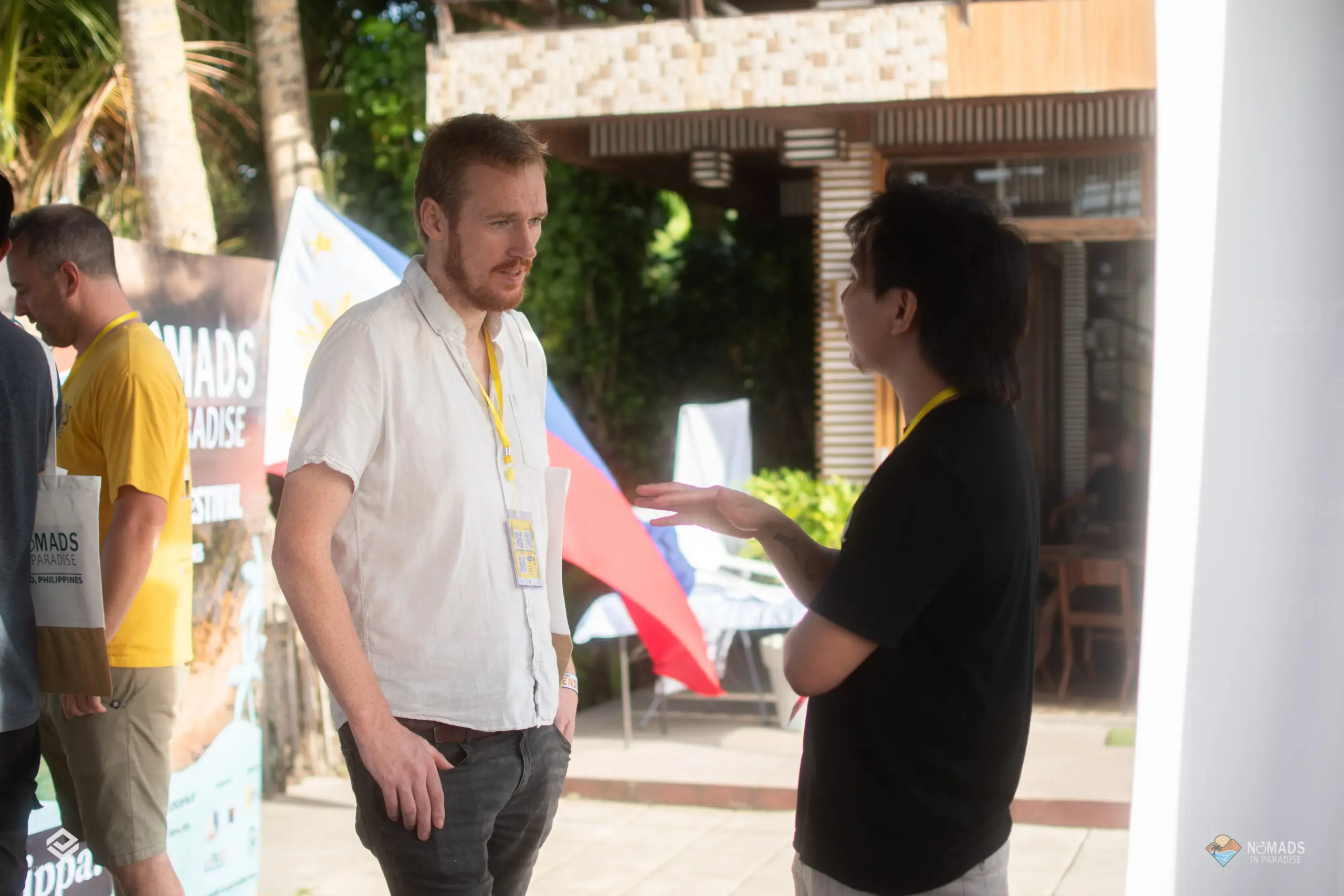 Two men networking at outdoor Nomads in Paradise festival with Philippine flag and palm trees