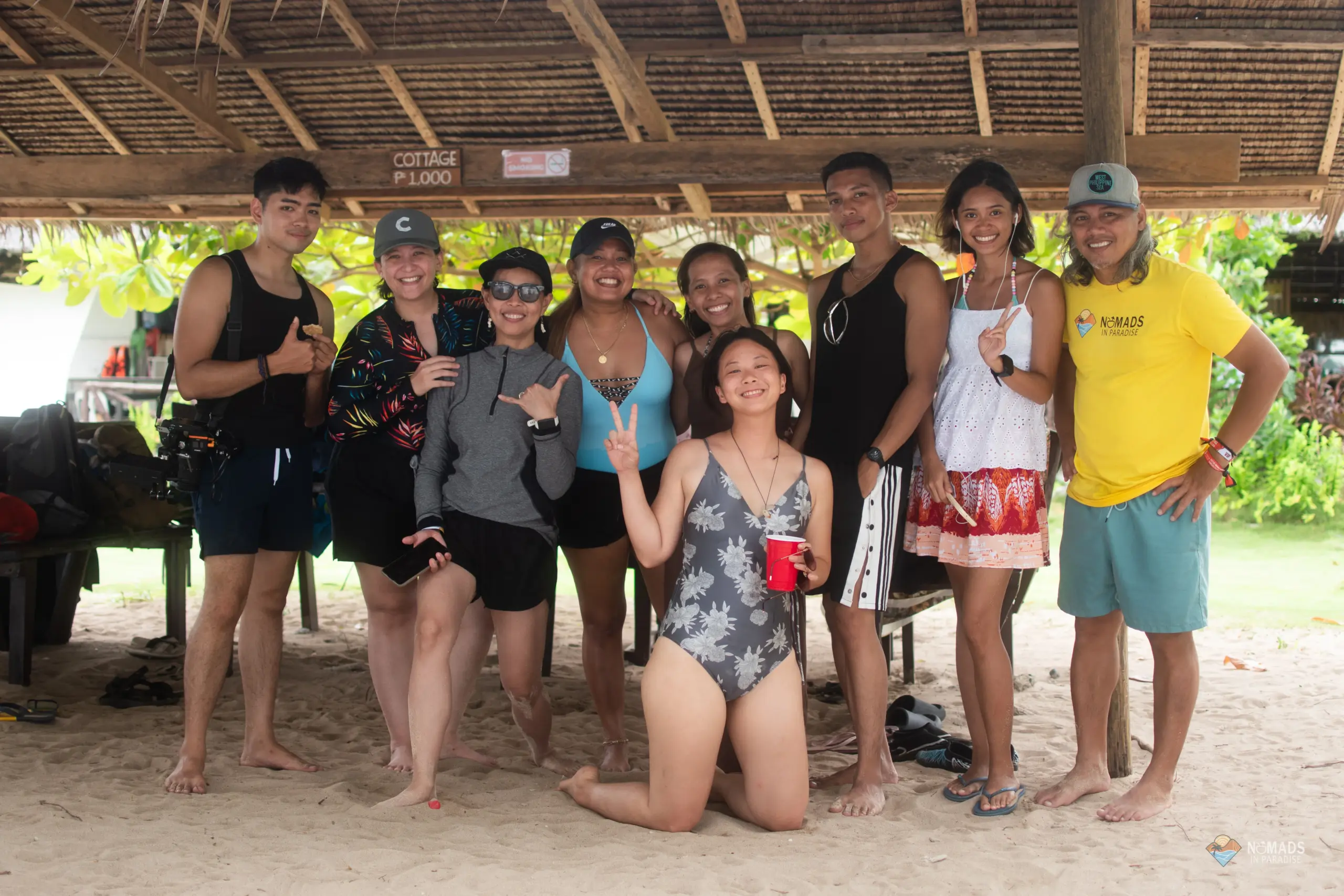 Nine happy Nomads in Paradise attendees posing under thatched beach cottage